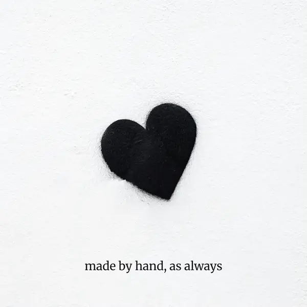 A black felt heart on a white wall, cropped square, with “made by hand, as always” set beneath it