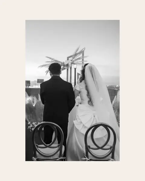 A black-and-white frame of a couple seen from behind at the altar, uncropped inside a 4:5 post on a warm cream background