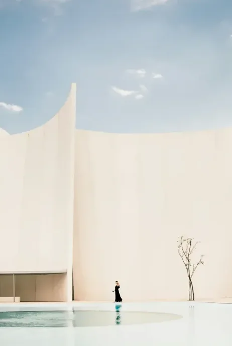 A solitary figure in a black dress walking by a reflecting pool in front of a curved white wall